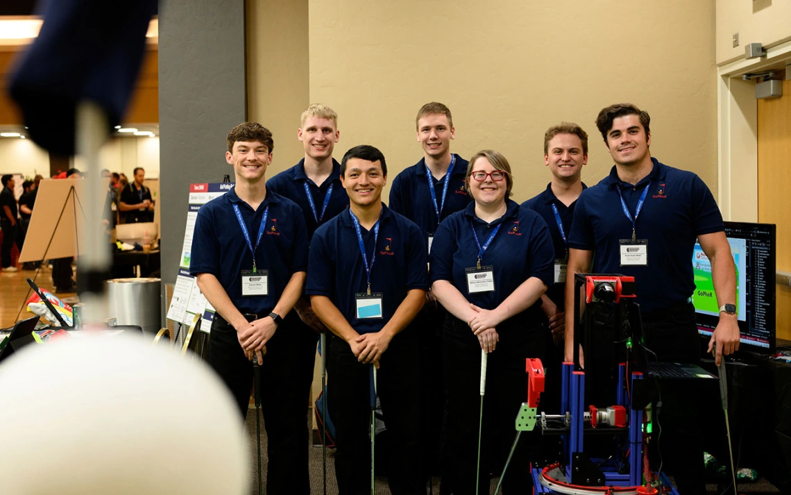 Six men and one women pose for a photo next to a robot with a golf club.